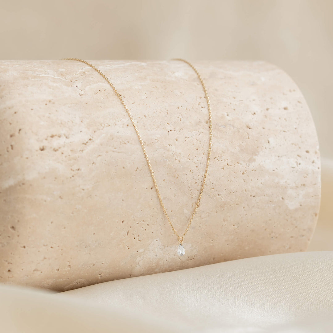 A delicate Gold necklace with a small crystal pendant drapes elegantly over beige stone. The soft, neutral background enhances its simplicity and elegance.