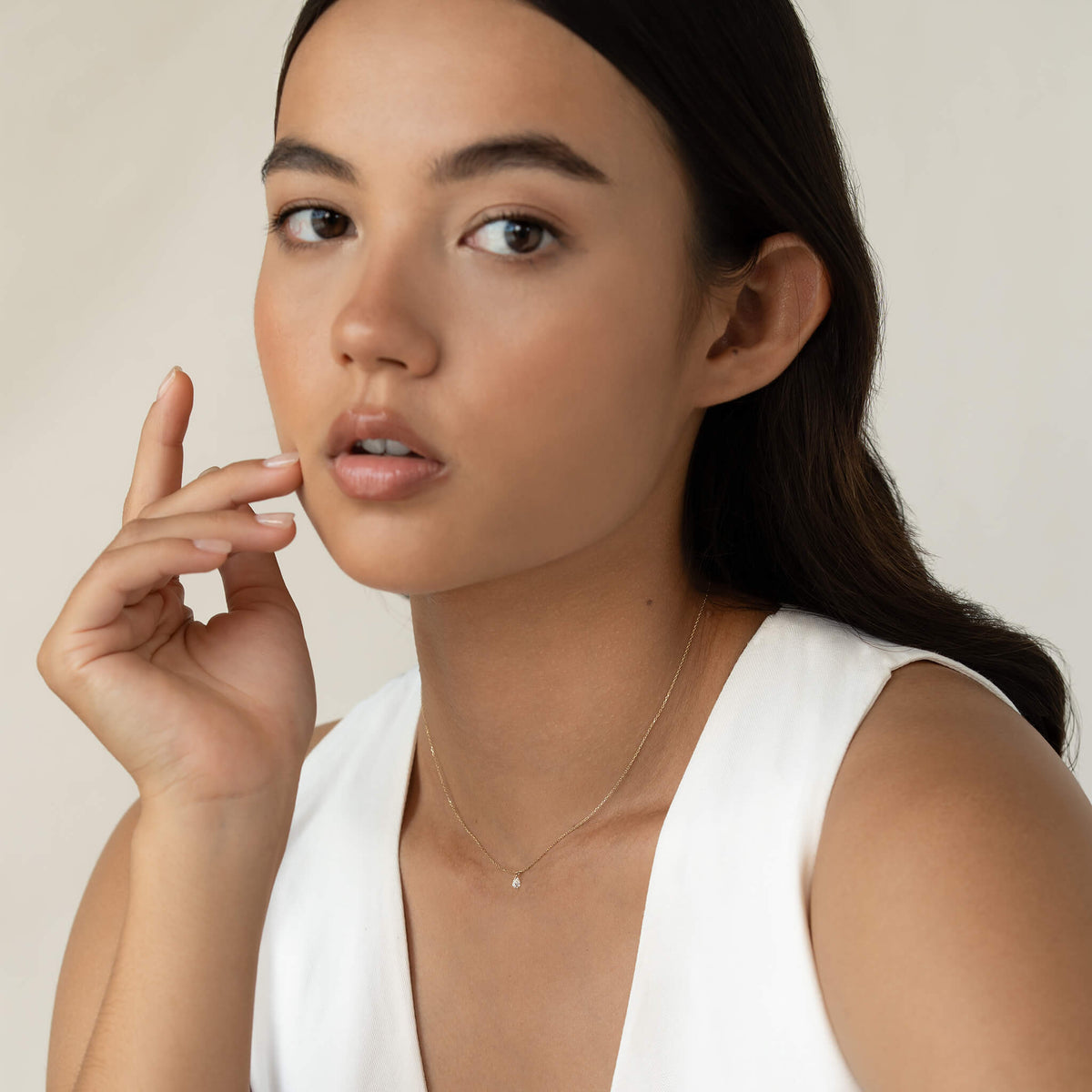 Young woman with long hair and neutral expression, wearing a white top and delicate Gold necklace, gently touches her face. Soft, neutral background.