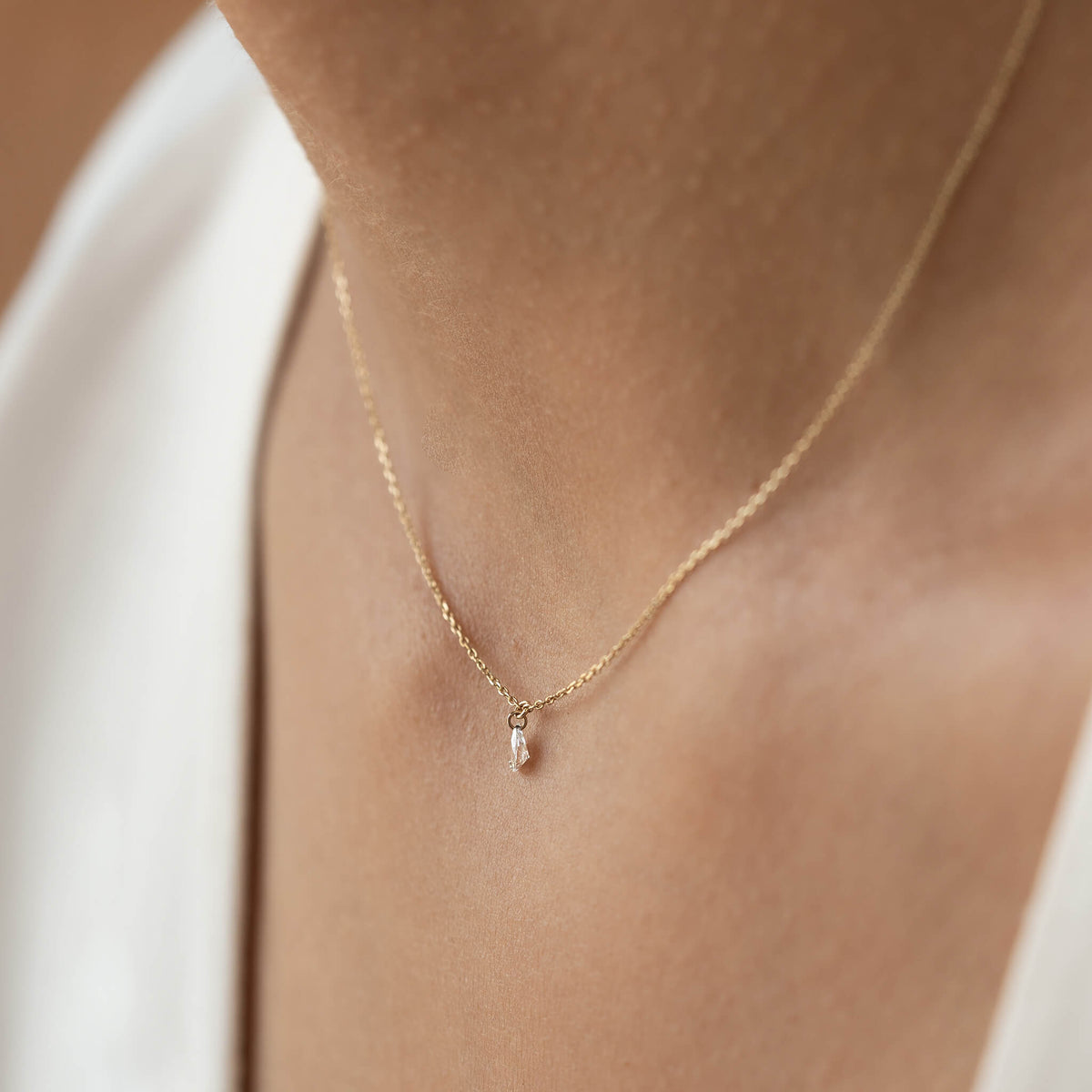 A delicate Gold chain necklace with a small teardrop pendant adorns a person's neck, set against the background of a white top, conveying elegance.