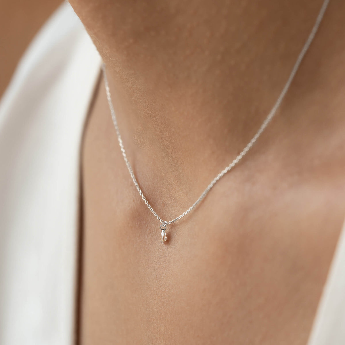 Close-up of a delicate White Gold chain necklace with a tiny teardrop pendant resting on a person's skin. The image conveys simplicity and elegance.