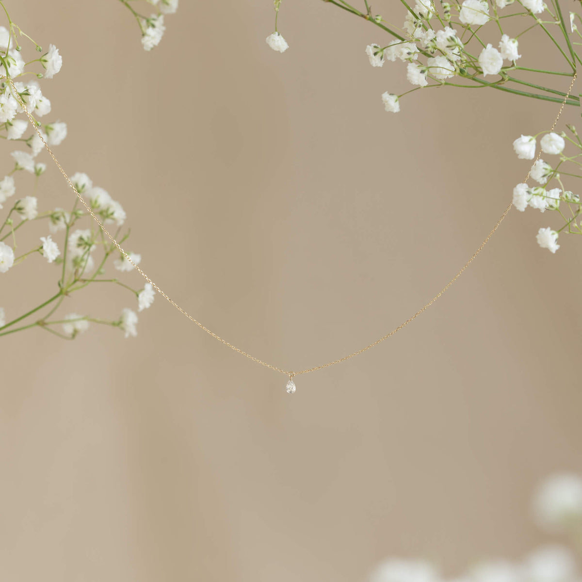 A delicate Gold chain with a small pendant is suspended between sprigs of white baby's breath flowers against a soft beige background, conveying elegance.