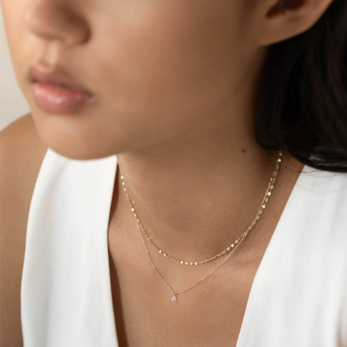 A close-up of a woman wearing two delicate Gold necklaces. One features small beads and the other has a tiny gem. She's dressed in a white top, conveying elegance.