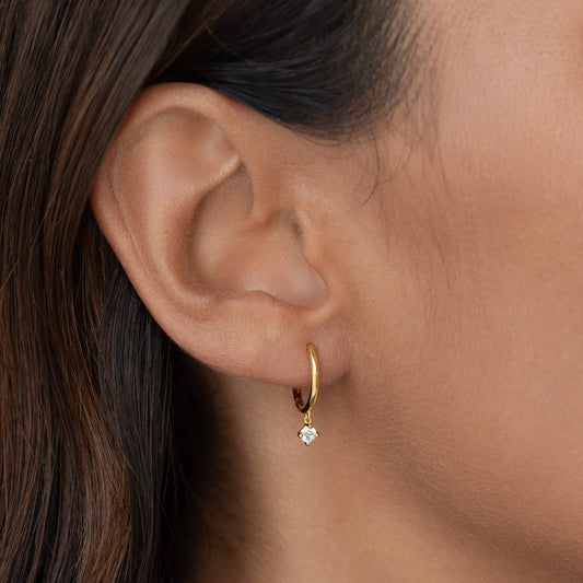 Close-up of a woman’s ear wearing a polished Gold Vermeil hoop earring with a small dangling clear gemstone.
