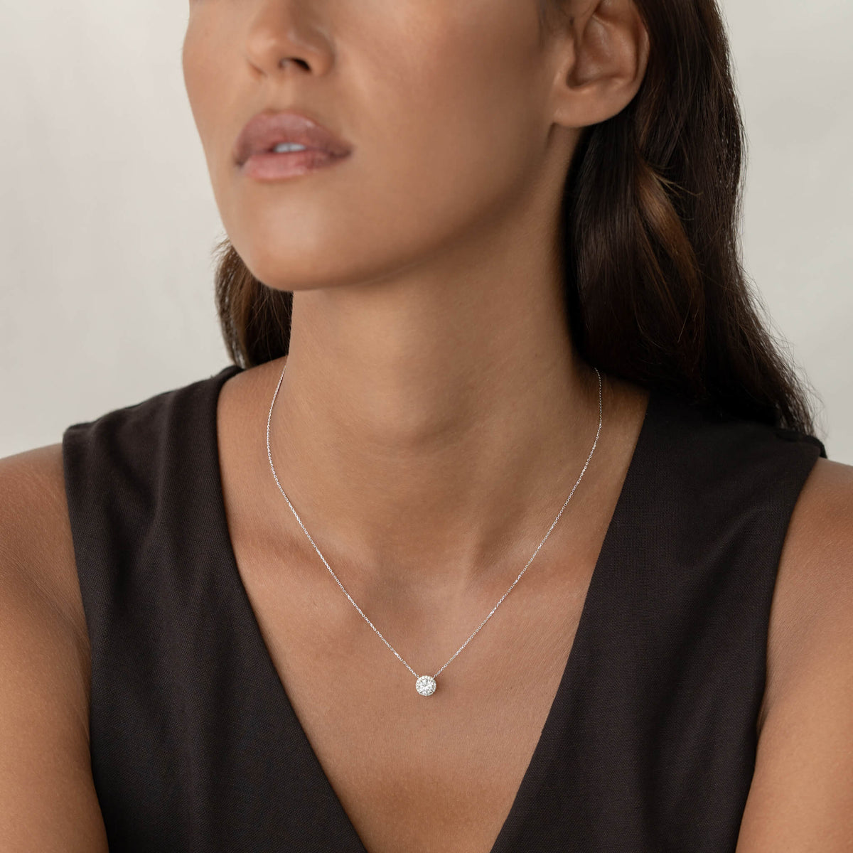 A close-up view of a person wearing a delicate White Gold chain necklace with a round brilliant-cut diamond halo pendant, worn against a simple dark-colored top.