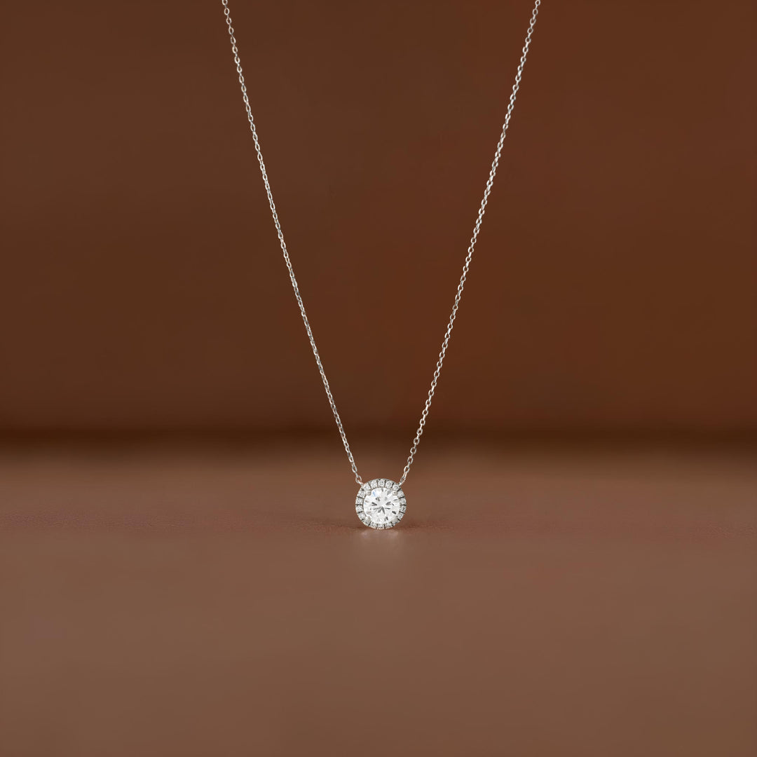 A close-up shot of a delicate White Gold necklace featuring a small, round brilliant-cut diamond halo pendant, suspended elegantly against a soft, brown-colored fabric background.