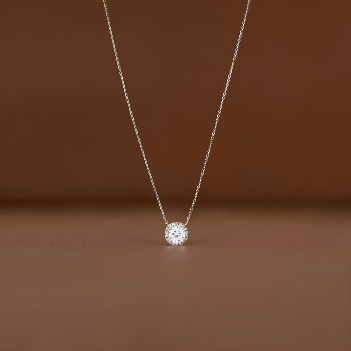 A close-up shot of a delicate White Gold necklace featuring a small, round brilliant-cut diamond halo pendant, suspended elegantly against a soft, brown-colored fabric background.