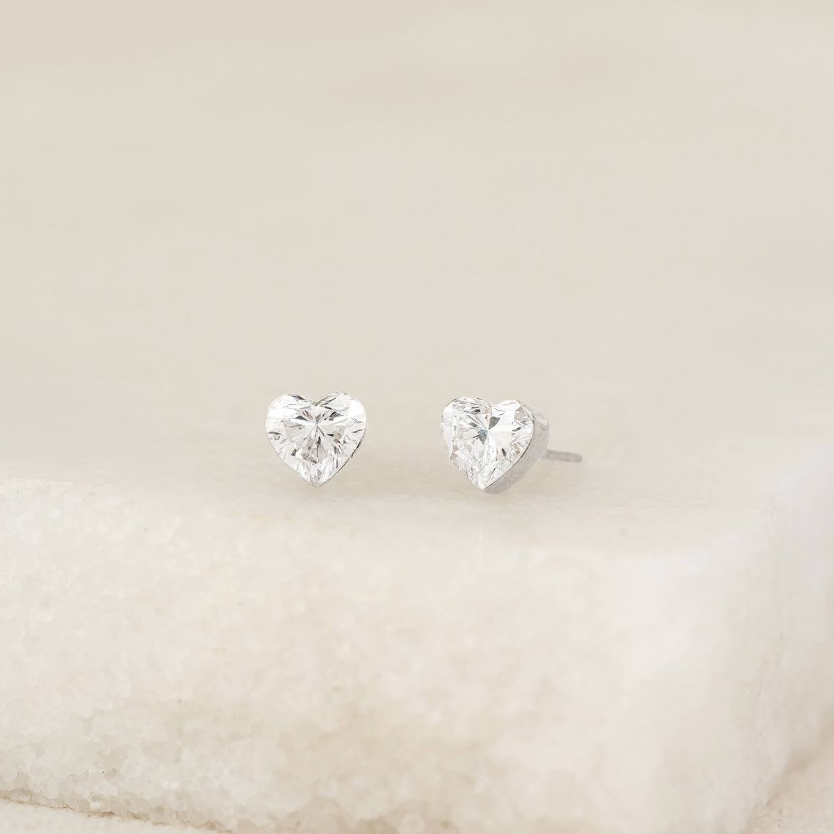 Pair of heart-shaped diamond stud earrings with White Gold settings, displayed upright on a soft, neutral textured surface against a minimal background.