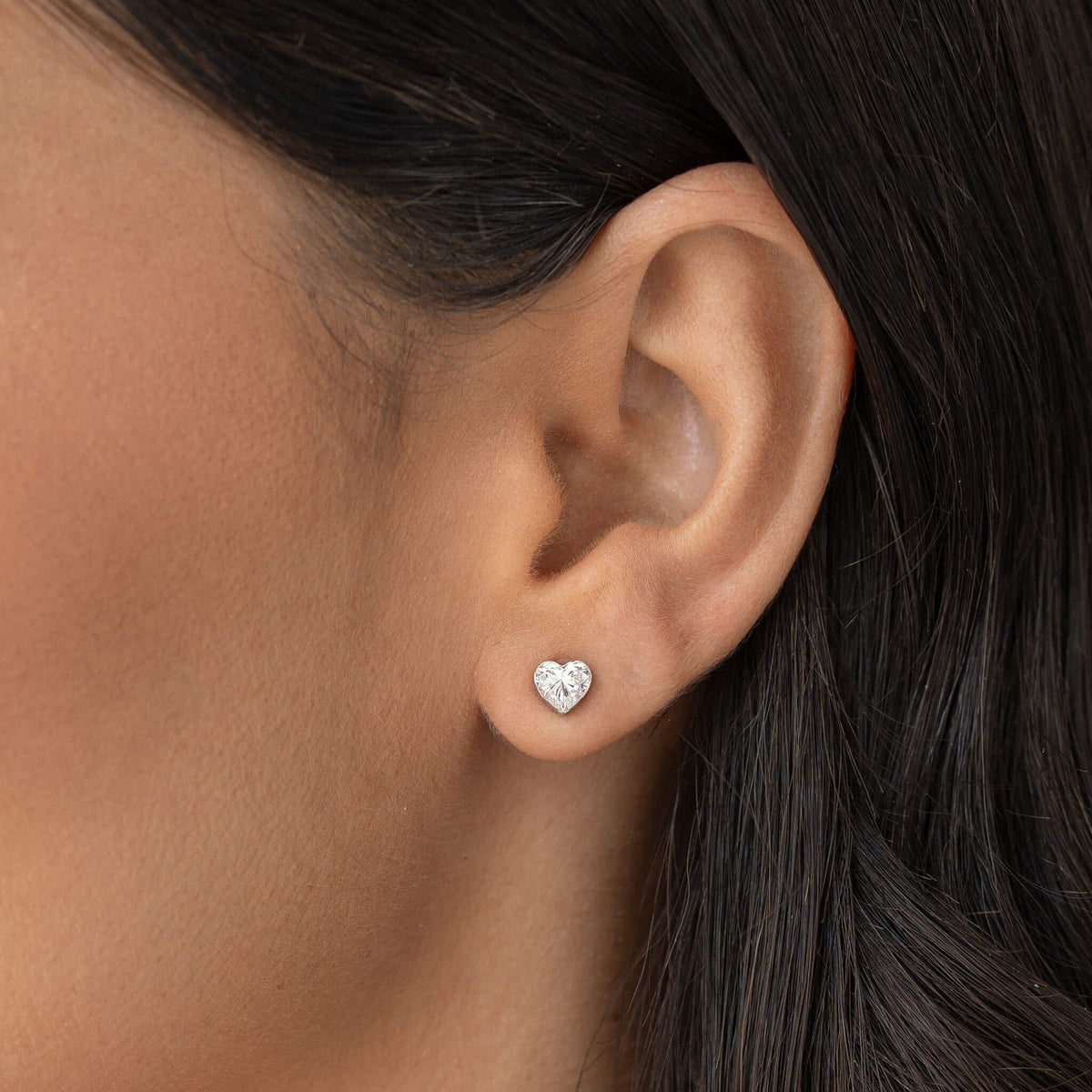 Close-up of a person’s ear wearing a small White Gold heart-shaped diamond stud earring, with smooth skin and dark hair tucked behind the ear.