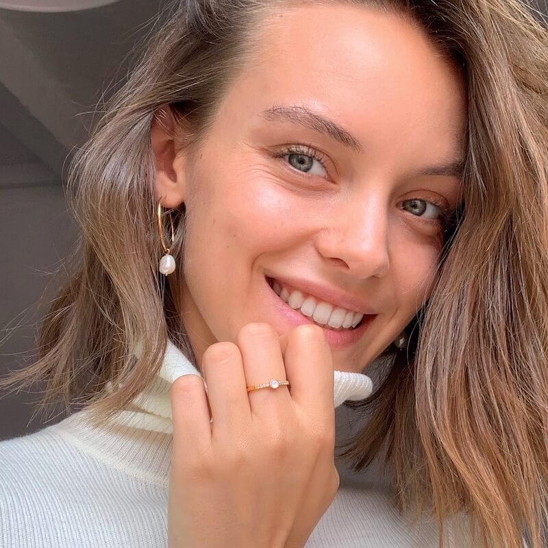 Close-up of a smiling woman with light brown hair wearing pearl drop earrings and a white turtleneck sweater indoors