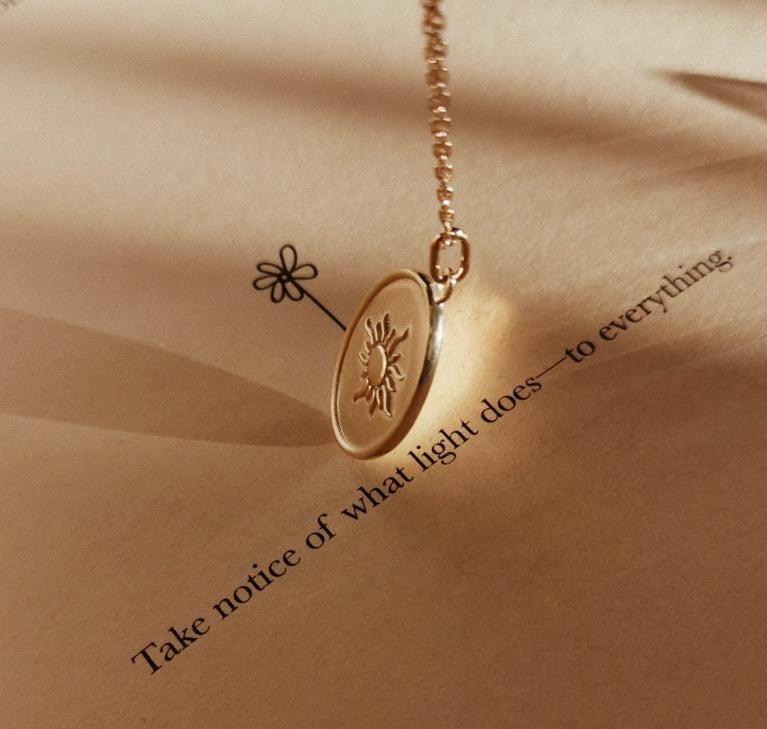 Close-up of a gold pendant necklace with sun design casting shadow on beige paper with printed inspirational text