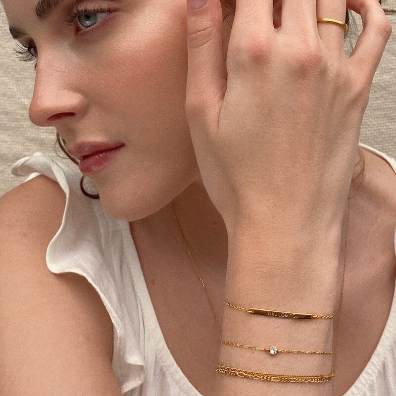 Close-up of a woman wearing layered delicate gold bracelets and a white ruffled sleeveless top against a neutral background