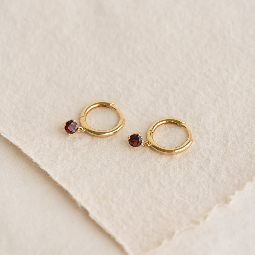 Close-up of January Birthstone Drop Huggie Earrings - Garnet with gold vermeil setting on a textured beige background