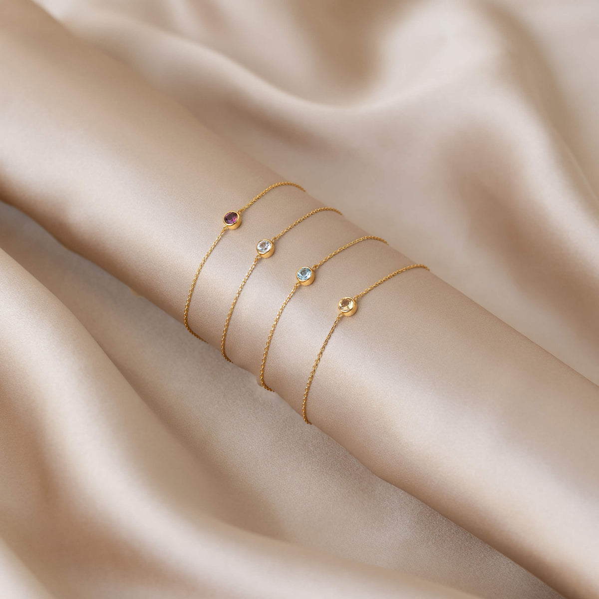 Four delicate Gold Vermeil bracelets with small round gemstones in different colors, elegantly arranged across soft champagne-colored satin fabric.