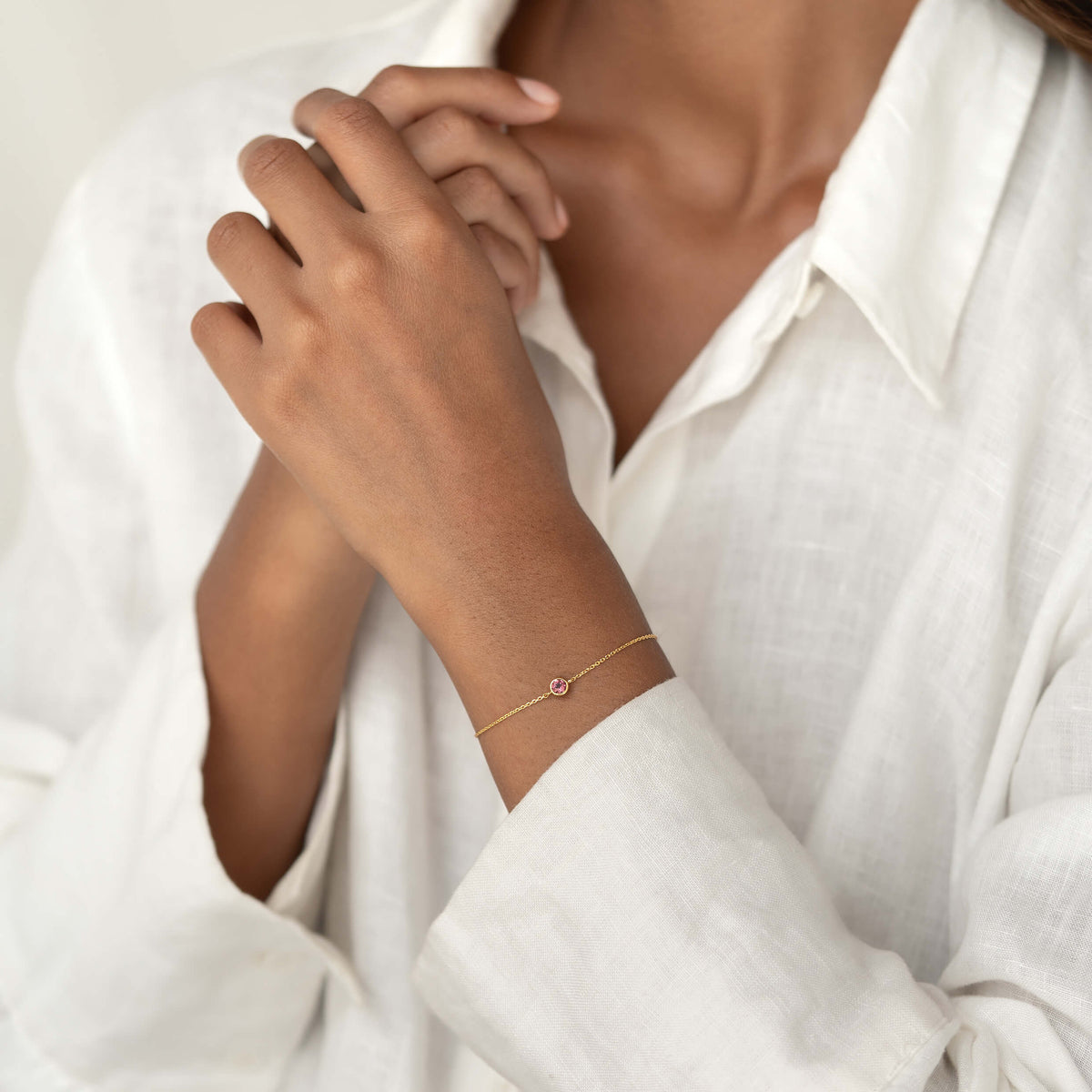 Close-up of a person wearing a white linen blouse, gently resting one hand while showcasing a delicate Gold Vermeil bracelet with a small Dark pink tourmaline gemstone centerpiece.
