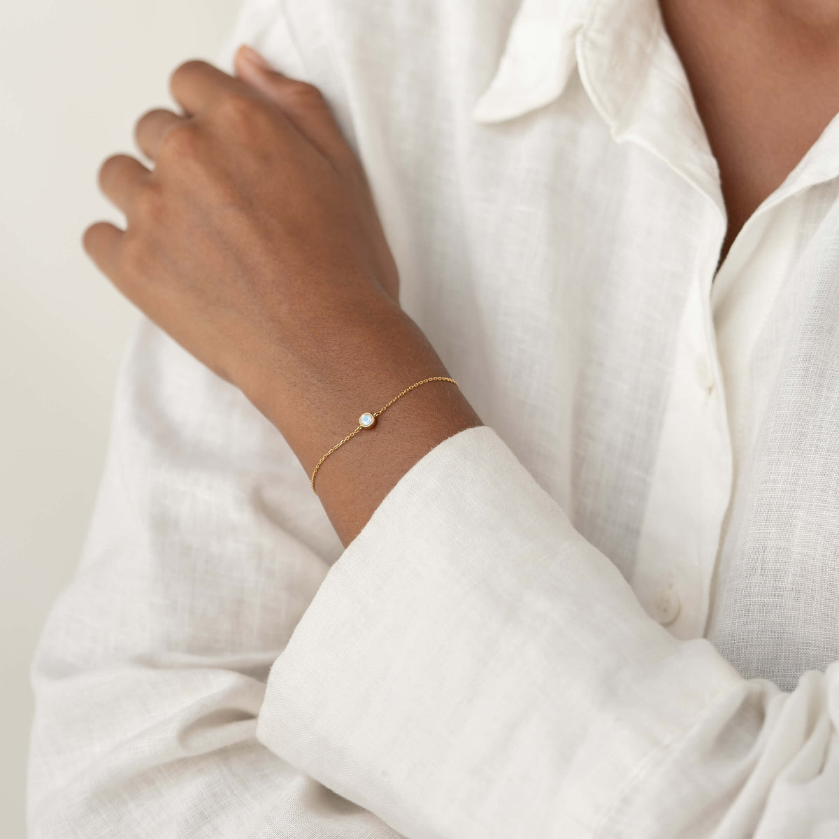 Close-up of a person wearing a white linen blouse, gently resting one hand on their shoulder while showcasing a delicate Gold Vermeil bracelet with a small Moonstone gemstone centerpiece.