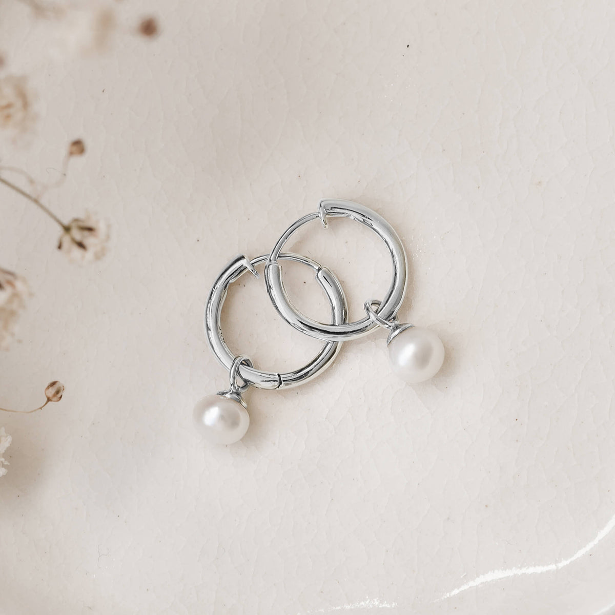 Silver hoop earrings with dangling pearls lie on a textured, light cream surface. Delicate dried flowers are visible in the top left corner.
