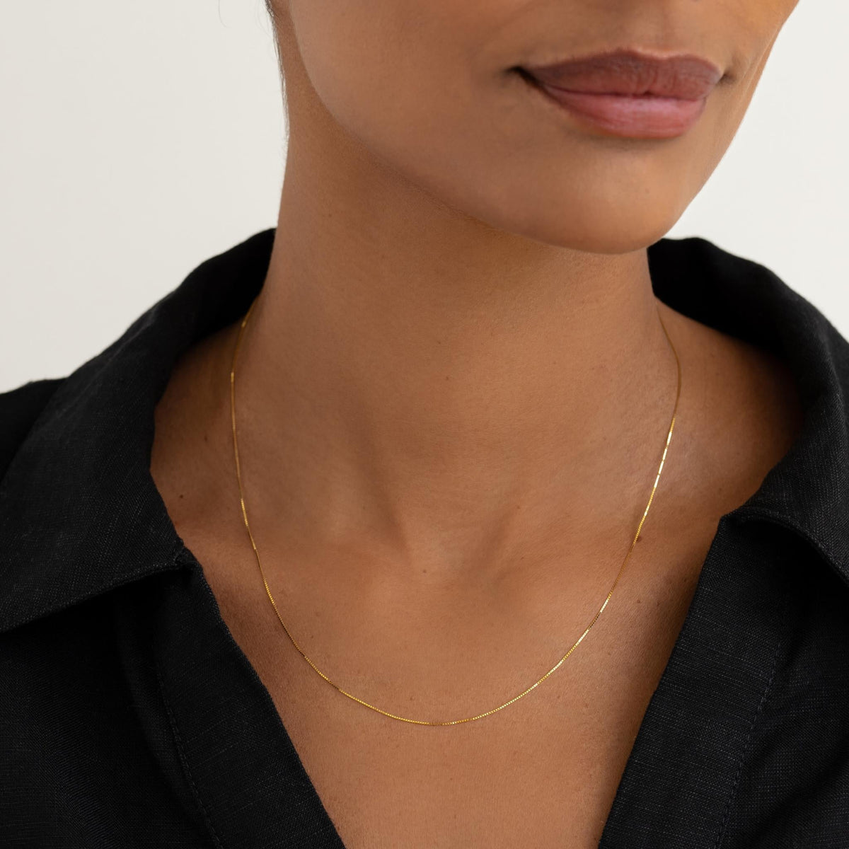 Close-up of a woman wearing a delicate 14k gold box chain necklace styled with a black collared shirt on a white background