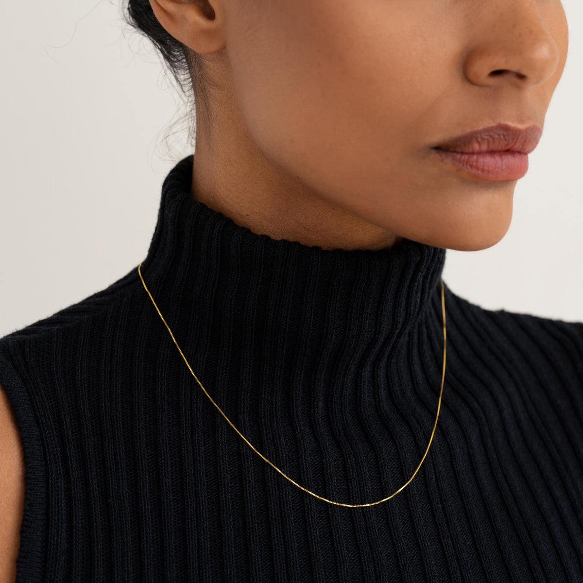 Close-up of woman wearing a delicate 14k gold box chain necklace with a black ribbed turtleneck sweater