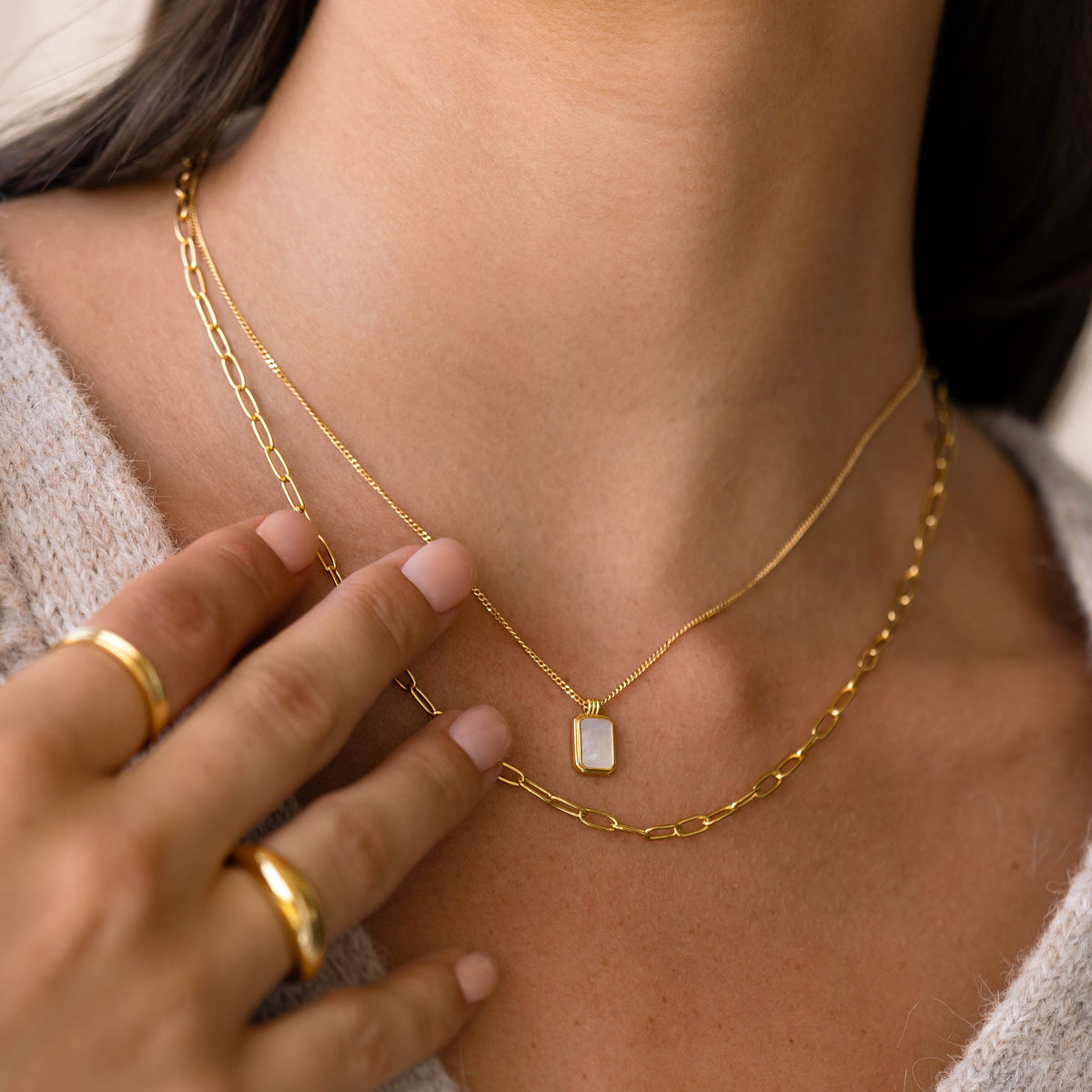 Close-up of a woman wearing the Mother of Pearl Tag Necklace - Iria layered with gold chains and rings on a beige sweater