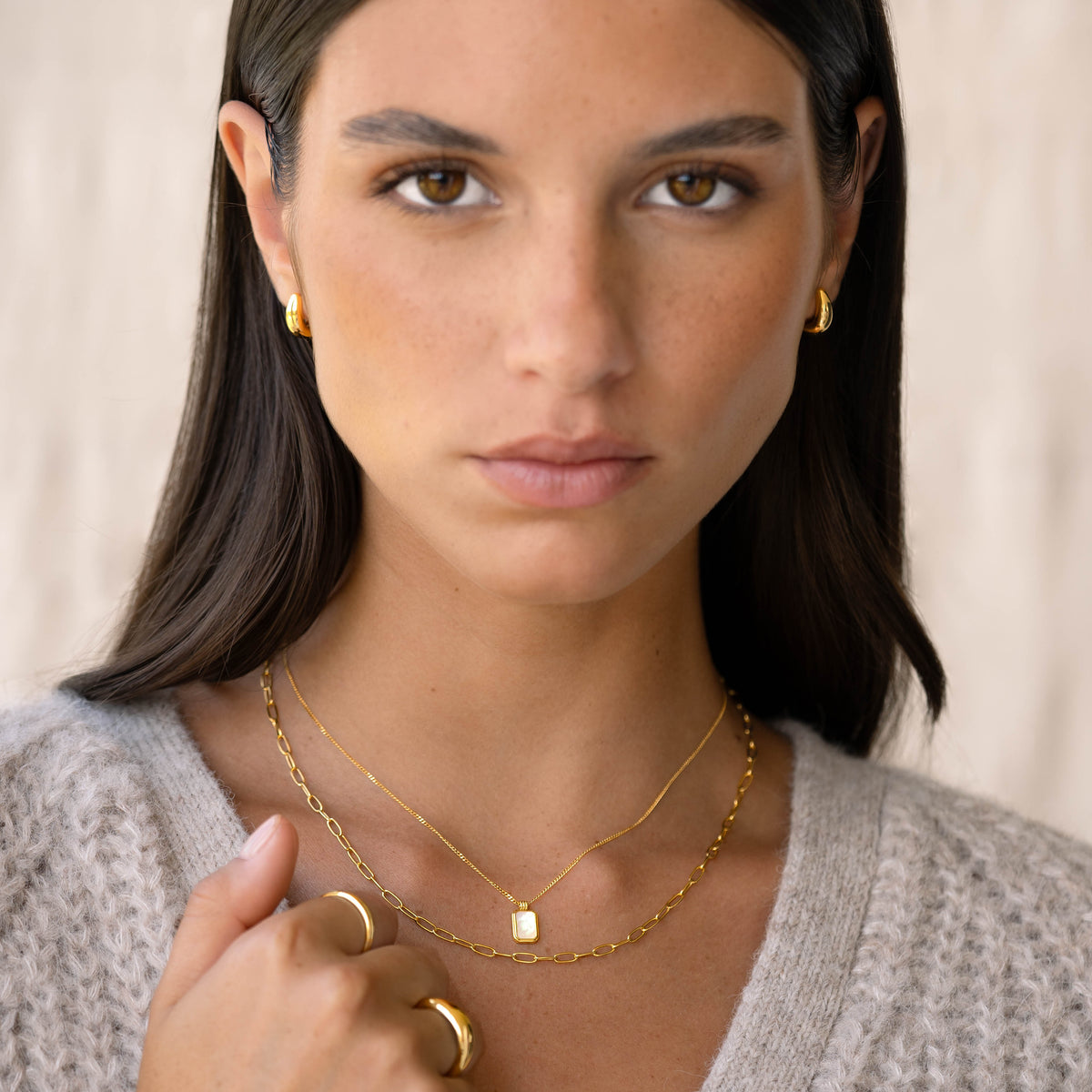 Close-up of a woman wearing Dome Huggie Earrings - Caja in gold vermeil, paired with gold necklaces and rings against a neutral background