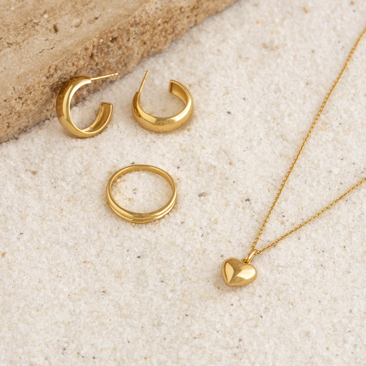 Close-up of gold vermeil Double Stacking Ring - Idalia with matching gold hoop earrings and heart pendant necklace on sandy surface