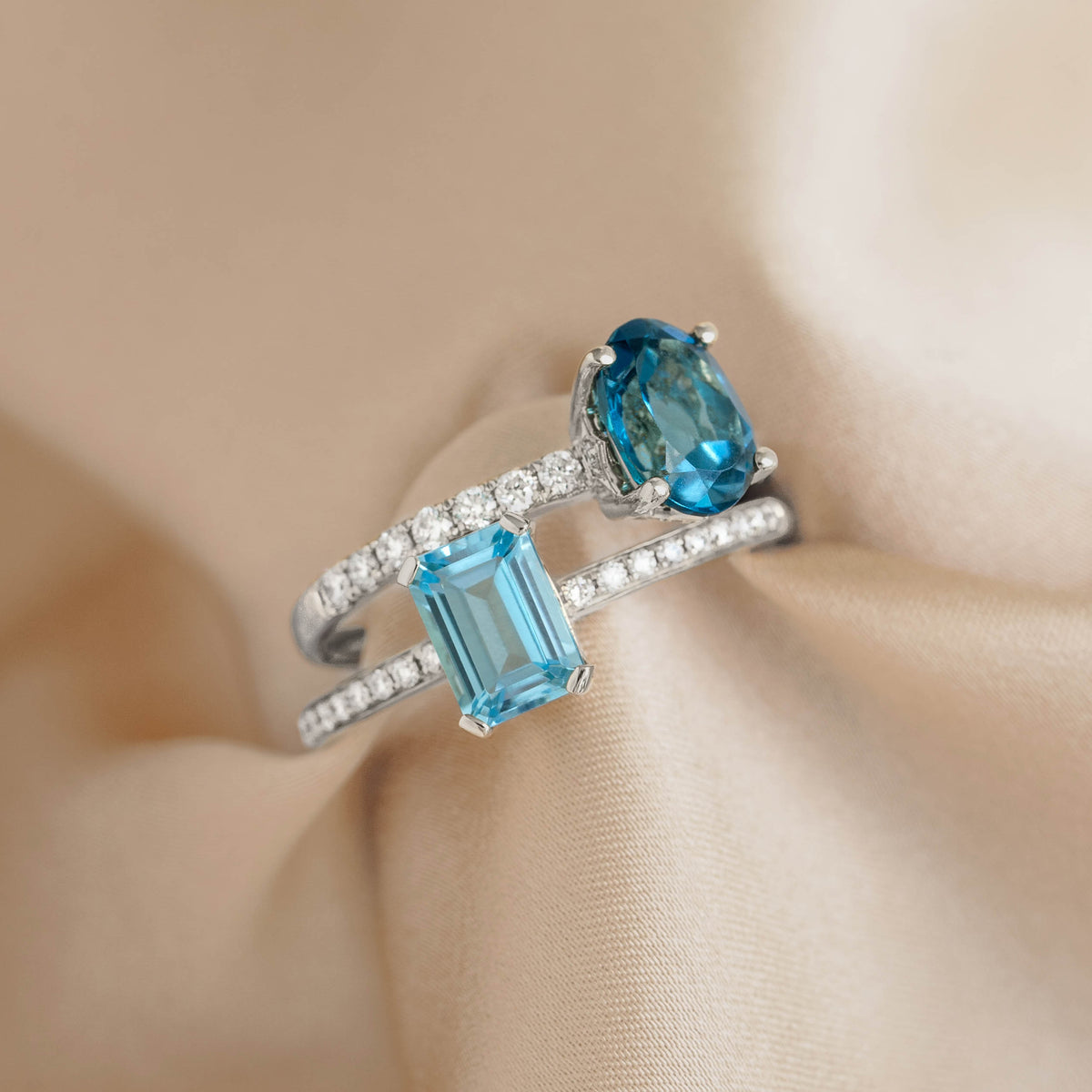 Close-up of Baguette Swiss Blue Topaz and Diamond Ring White Gold - Safiya with two blue stones and pavé diamond bands on beige fabric