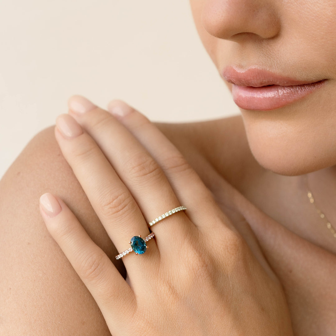 Close-up of a hand wearing the Oval London Blue Topaz and Diamond Ring Rose Gold - Minette on a neutral background
