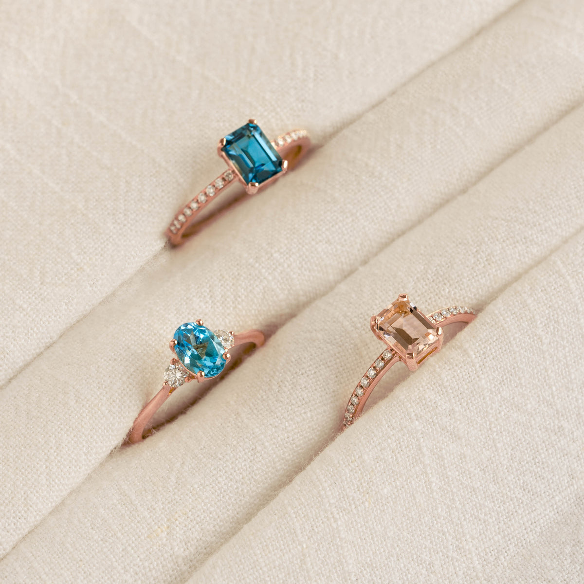 Close-up of three rose gold rings with baguette and oval gemstones on a beige fabric background featuring Baguette Morganite and Diamond Ring Rose Gold - Safiya