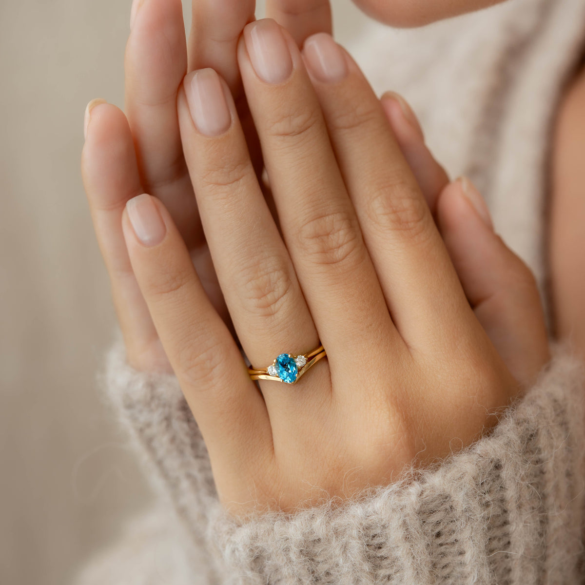Close-up of a hand wearing the Swiss Blue Topaz and Diamond Ring Rose Gold - Kindra with a cozy beige sweater background