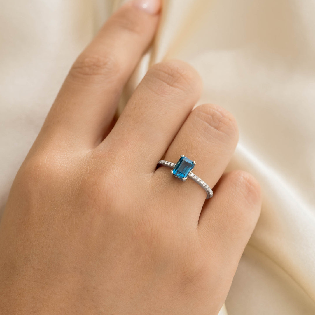 Close-up of a hand wearing the Baguette London Blue Topaz and Diamond Ring White Gold - Safiya on a soft beige fabric background