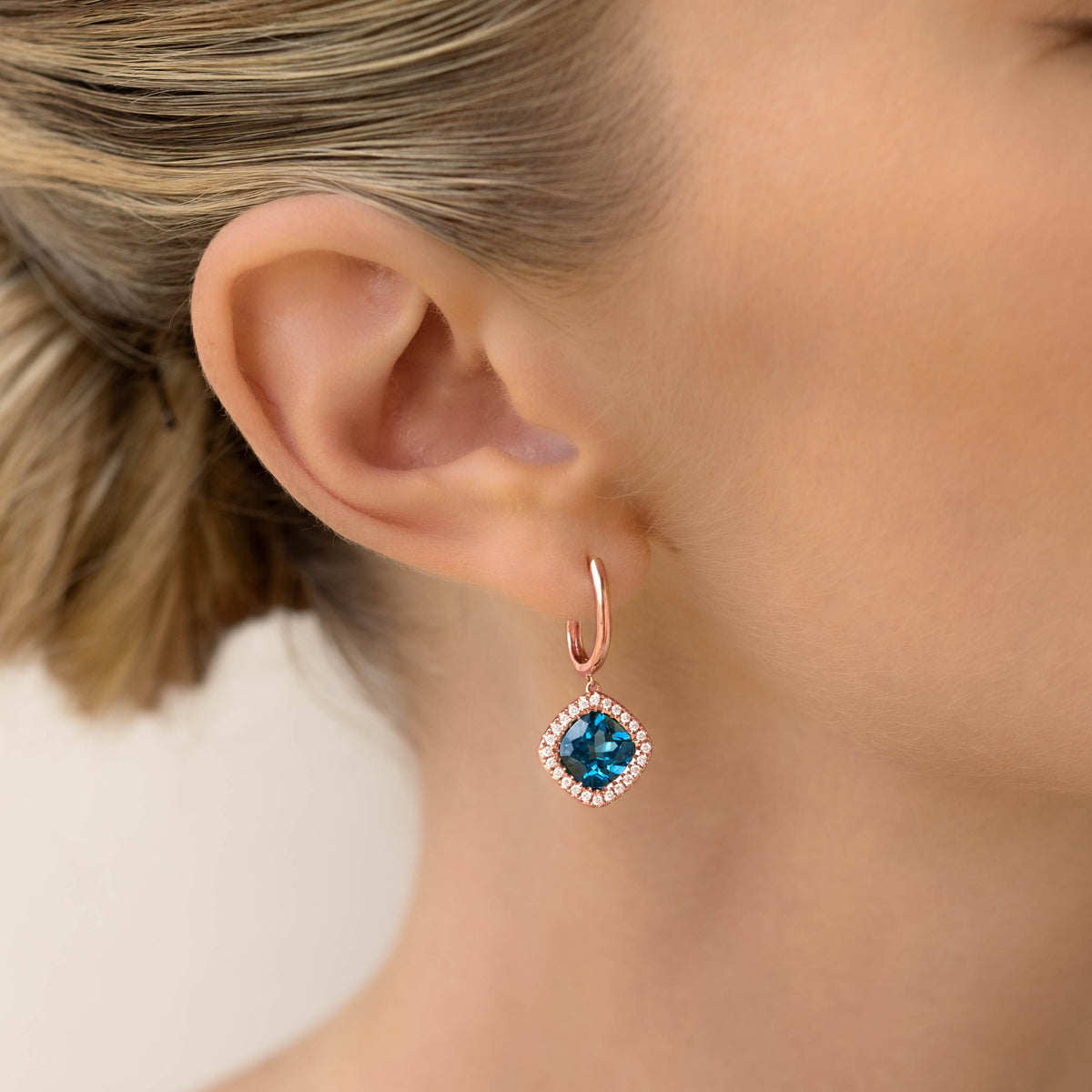 Close-up of a woman wearing London Blue Topaz and Diamond Halo Drop Earrings Rose Gold - Elvira on neutral background