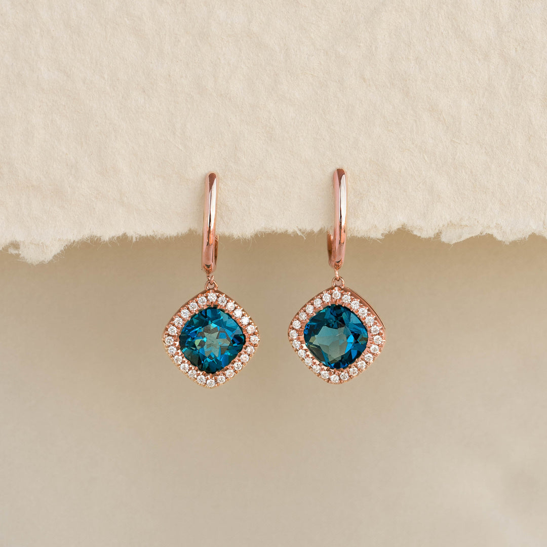 Close-up of London Blue Topaz and Diamond Halo Drop Earrings Rose Gold - Elvira against a neutral beige background