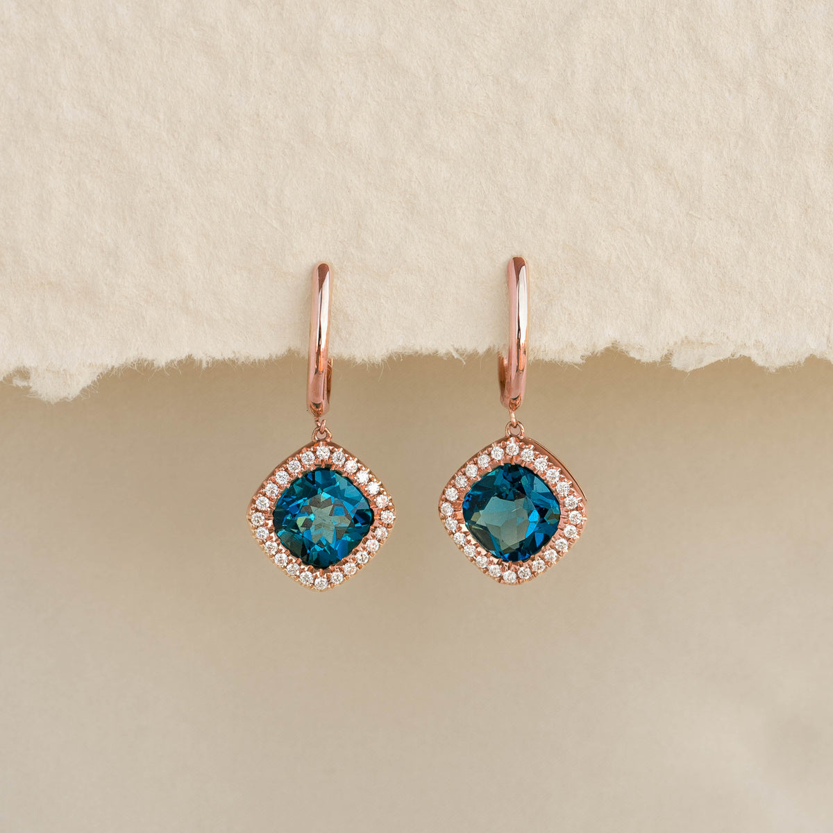 Close-up of London Blue Topaz and Diamond Halo Drop Earrings Rose Gold - Elvira against a neutral beige background