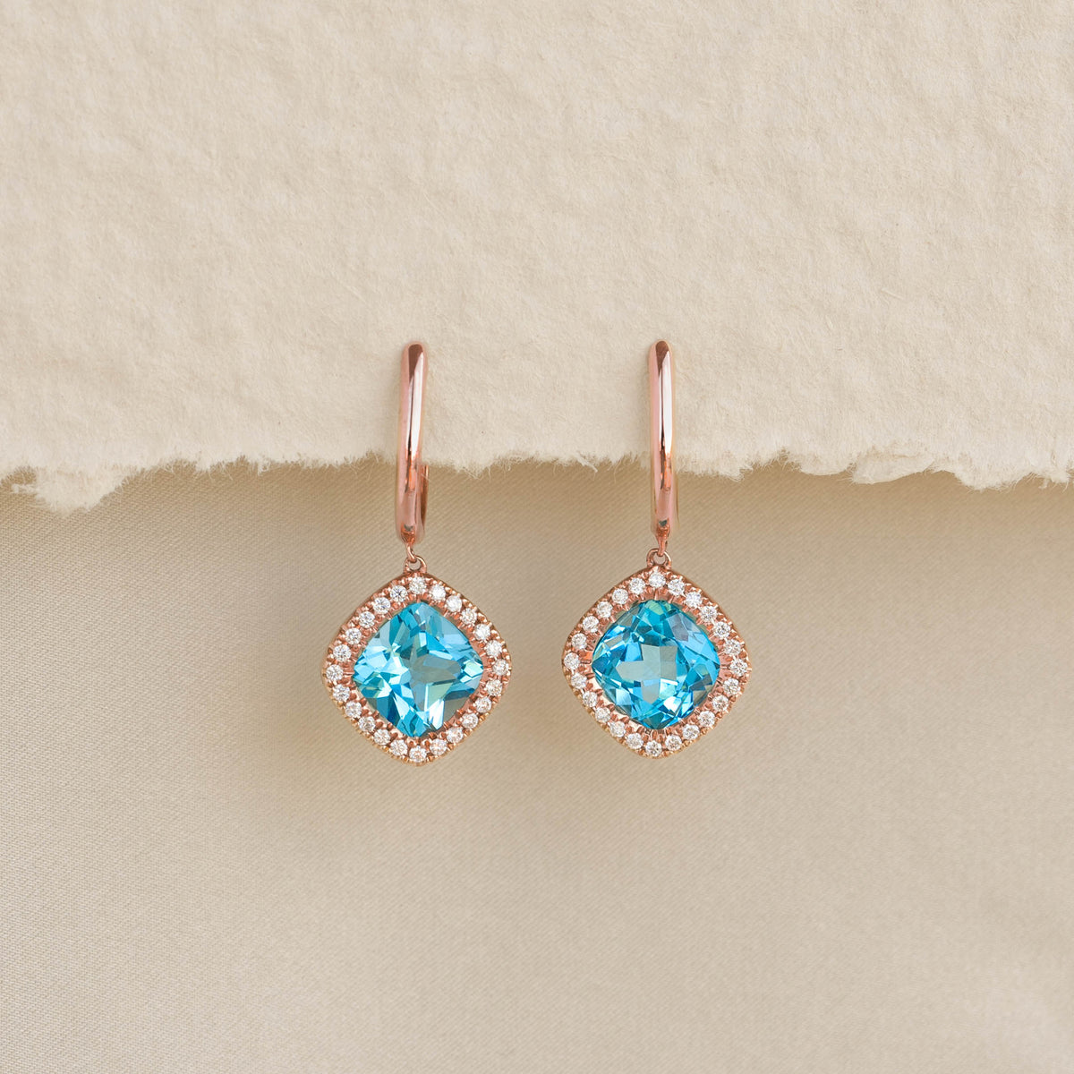 Swiss Blue Topaz and Diamond Halo Drop Earrings Rose Gold - Elvira with vibrant blue stones and sparkling diamond halo on beige background