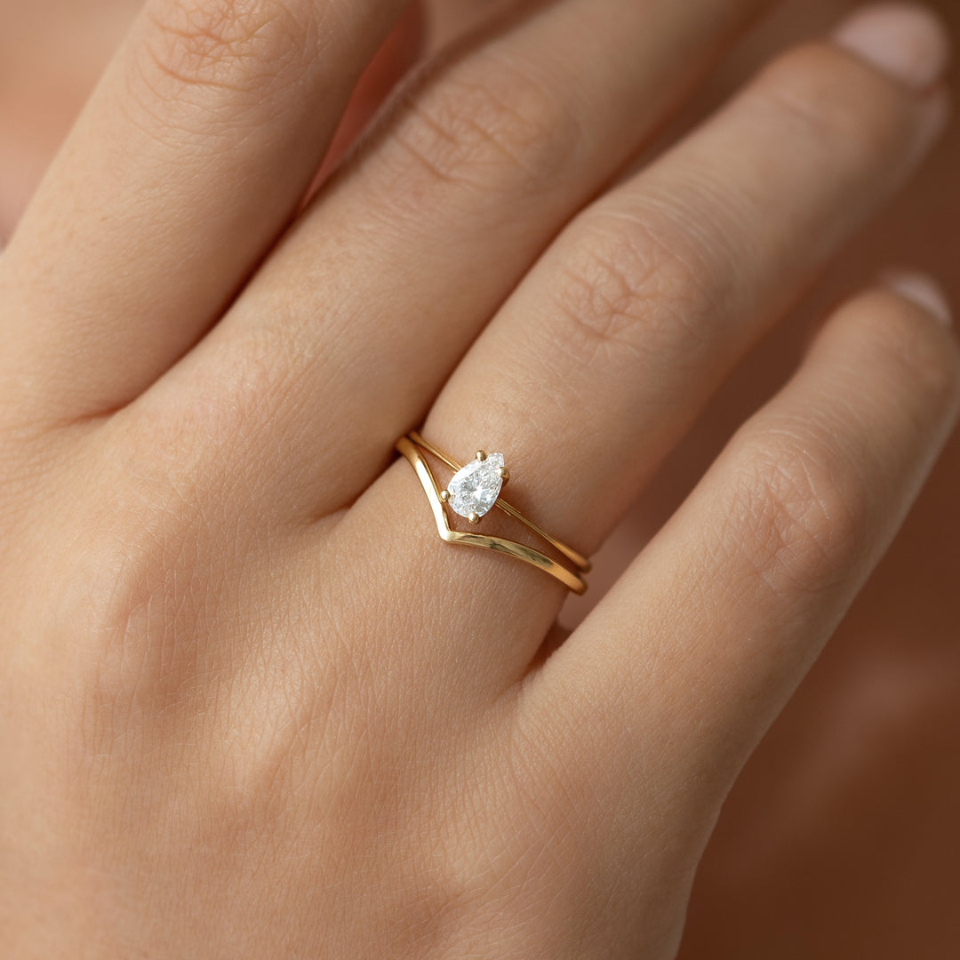 Close-up of a hand wearing the V Ring 14k Gold - Malva with a pear-shaped diamond and chevron band on a neutral background
