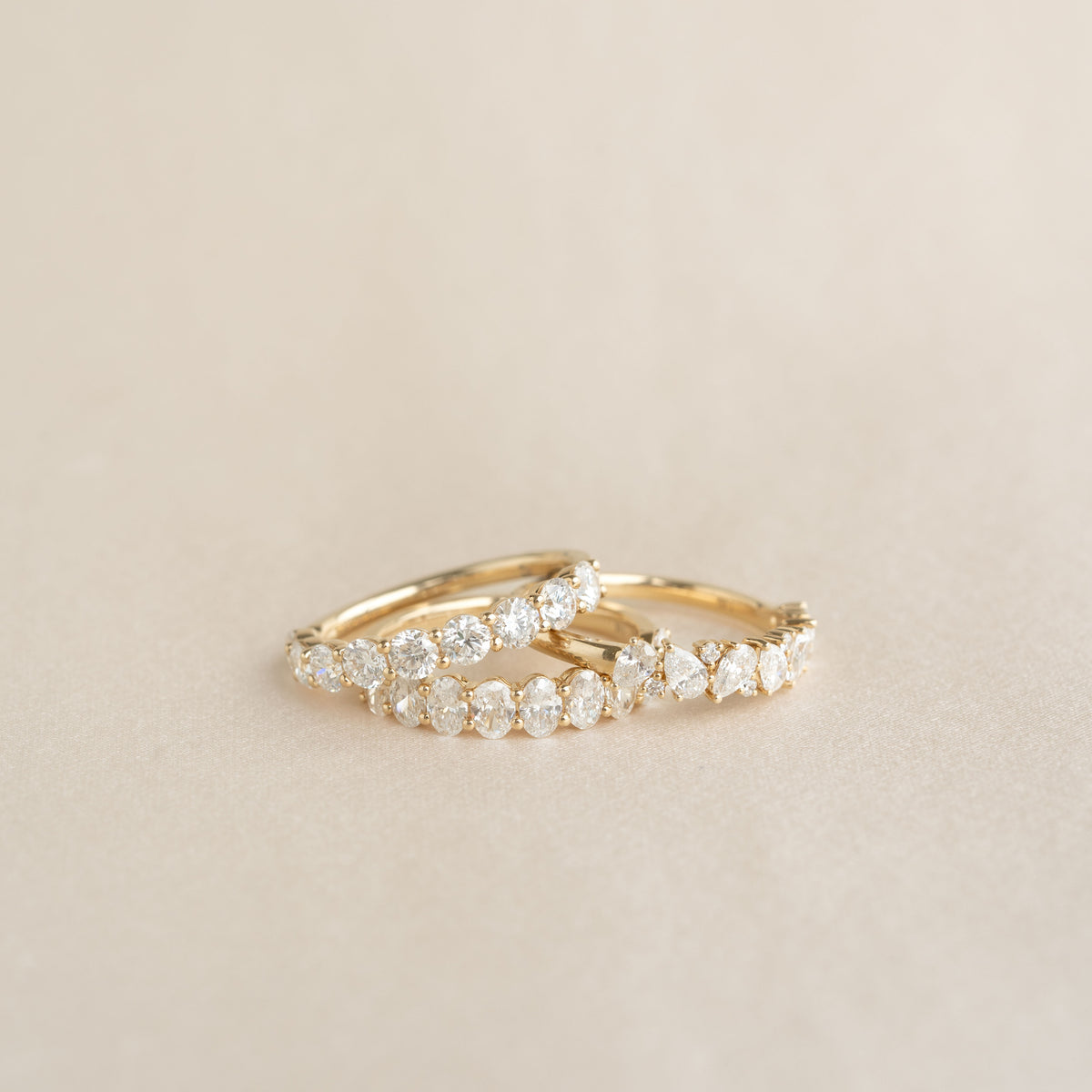Three Yellow Gold rings with diamond round cut, oval cut and pear-shaped settings are displayed on a soft beige surface.