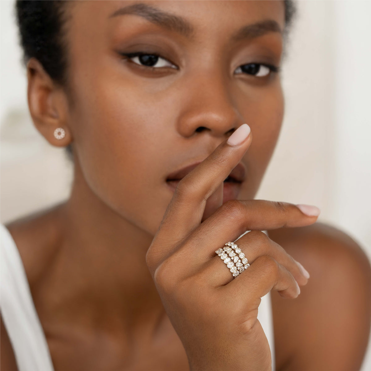 A close-up portrait of a model wearing a multi-row diamond ring, with her hand gently raised near her face, highlighting the stacked bands of round diamonds and a small stud earring against a soft neutral background.
