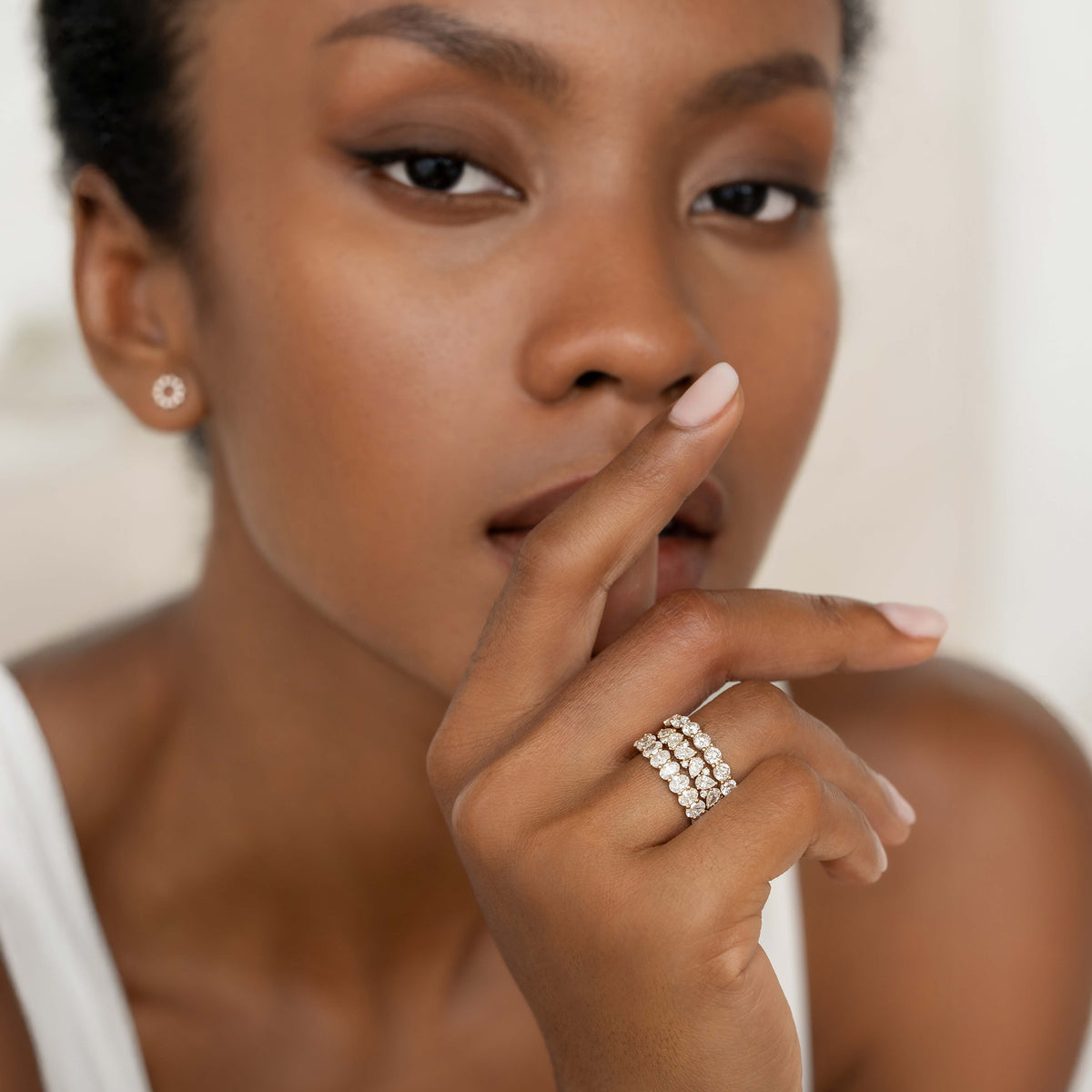 A close-up portrait of a model wearing a multi-row diamond ring, with her hand gently raised near her face, highlighting the stacked bands of round diamonds and a small stud earring against a soft neutral background.