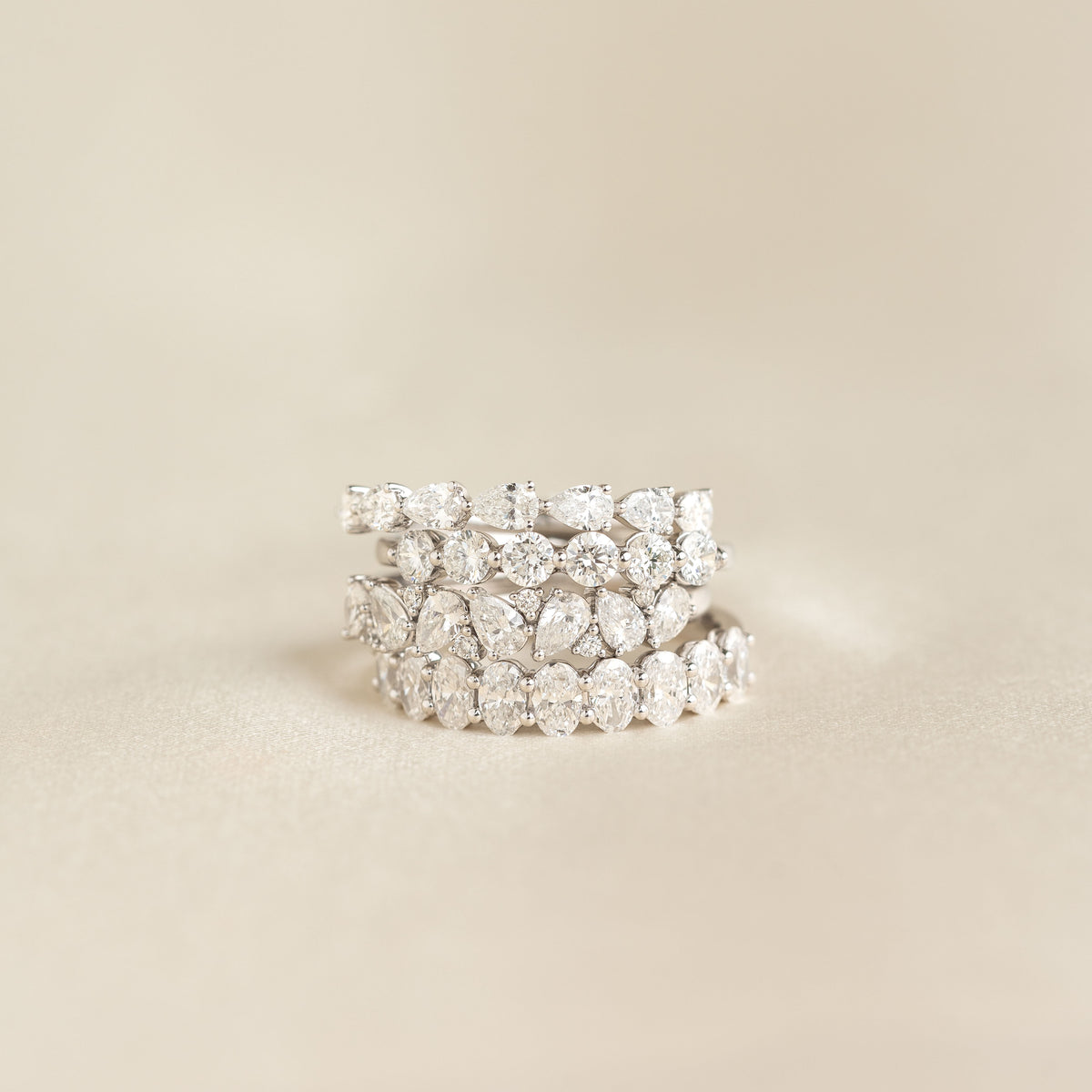 A stack of White Gold rings set with diamonds, featuring alternating pear-shaped and round stones, arranged in layered bands against a soft beige background.