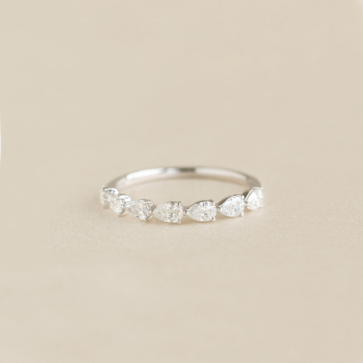 A delicate White Gold ring featuring a row of alternating pear cut diamonds set along the band, displayed on a soft beige background.