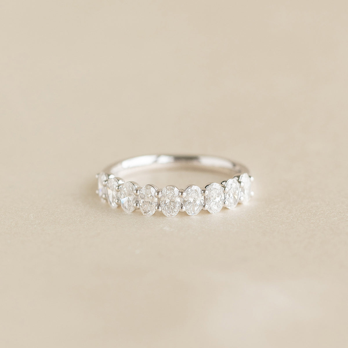 A delicate White Gold ring featuring a row of alternating oval cut diamonds set along the band, displayed on a soft beige background.