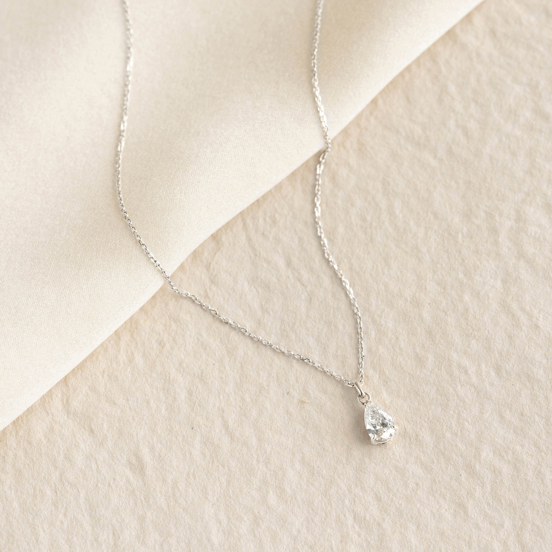 Pear Cut Diamond Pendant Necklace White Gold - Oline with a delicate chain and pear-shaped diamond on beige textured background