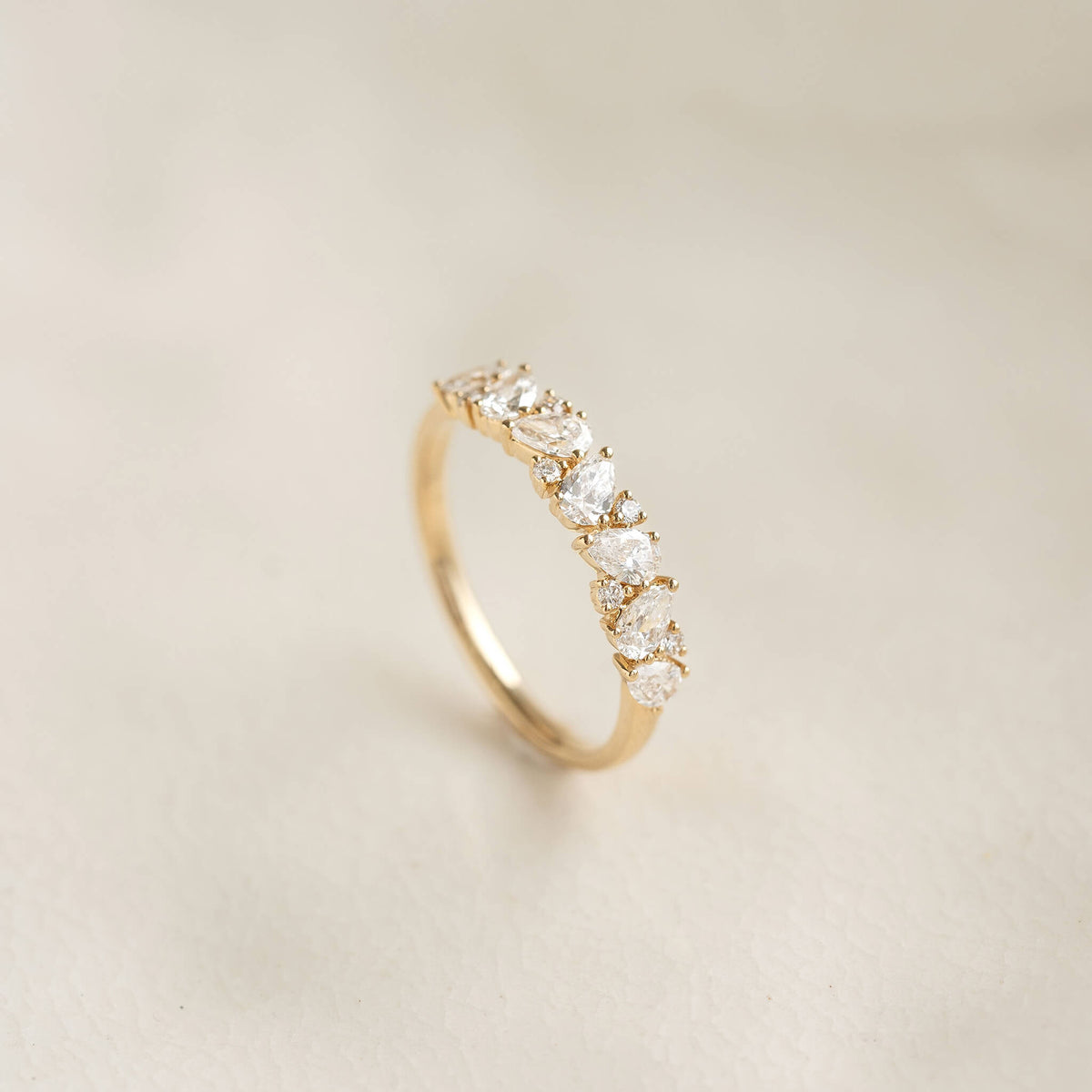 A delicate Yellow Gold ring displayed upright, featuring a row of alternating pear-shaped and round diamonds set against a soft beige background.