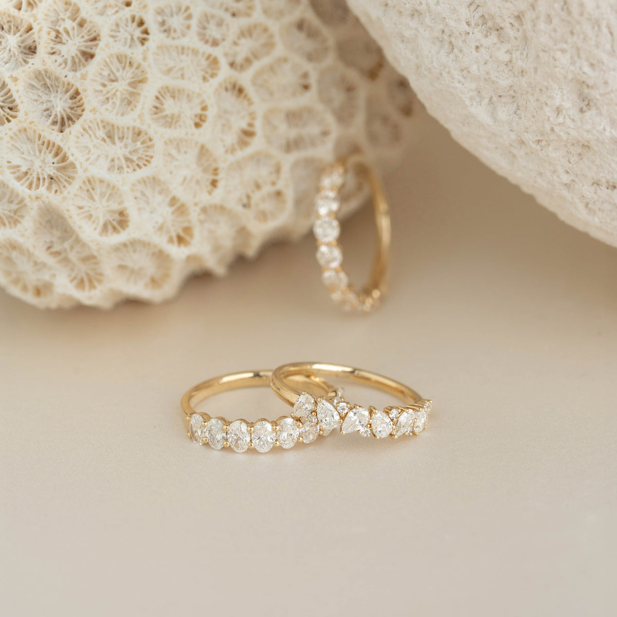 Three delicate Yellow Gold rings with diamond settings are displayed on a soft beige surface beside textured white coral.