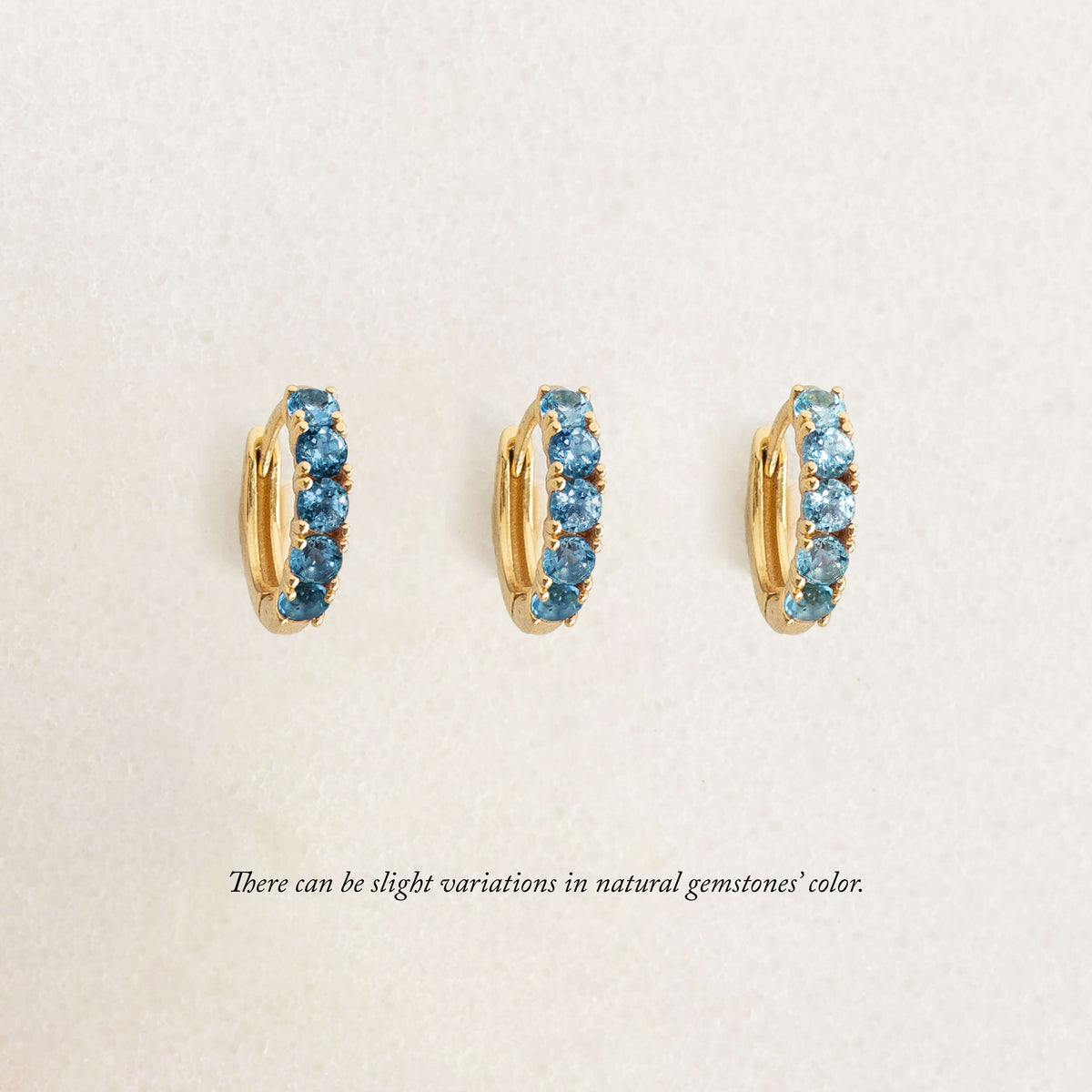 Three gold hoop earrings set with blue gemstones are displayed upright in a row on a light background. Text below notes slight color variations in natural gemstones.