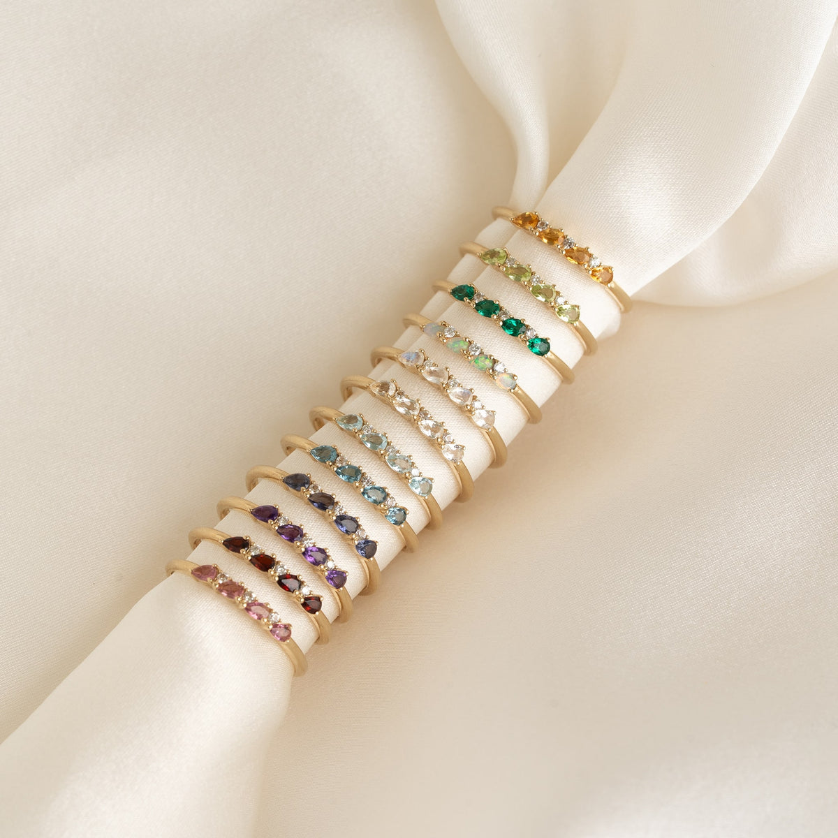 Close-up of May Birthstone Pear Ring 14k Gold - Emerald and Diamond styled with other gemstone rings on cream fabric background