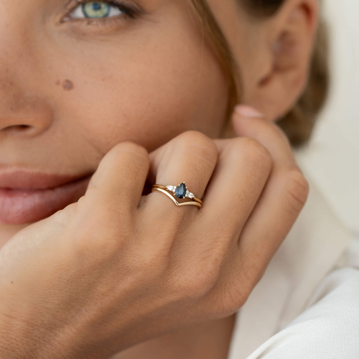 A close-up photograph shows a person resting their chin gently on their hand, showcasing a stacked Gold ring set on their ring finger.