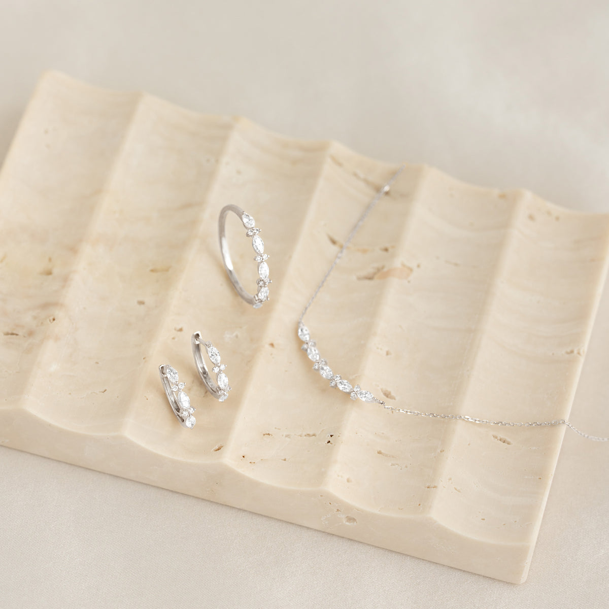 Marquise and Round Diamond Huggie Earrings White Gold - Zoya displayed on a textured beige tray with matching diamond ring and necklace