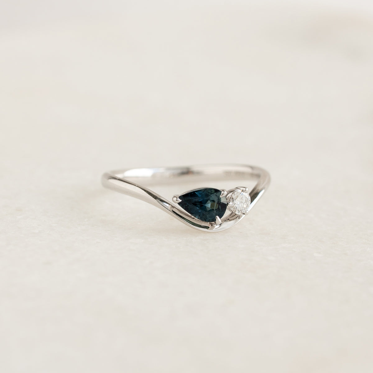 The White Gold ring features a sleek, asymmetrical wavy band that dips gracefully down to secure a horizontally-set, pear-cut deep teal sapphire.