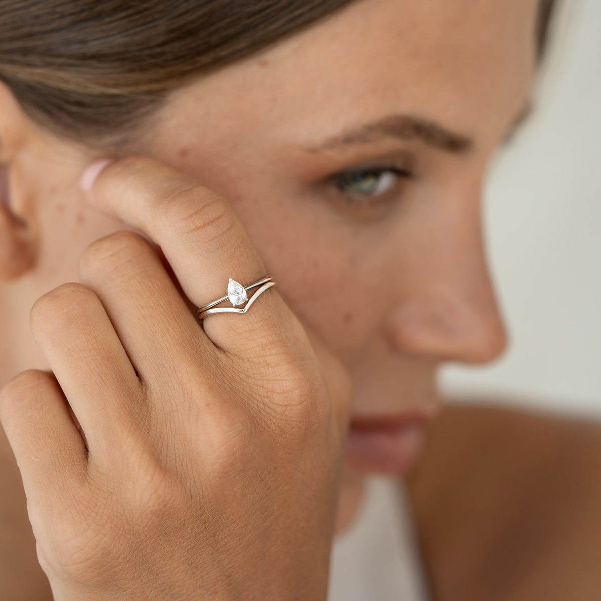 Close-up of a woman's face in soft focus, highlighting a pear-shaped diamond White Gold ring on her finger. The tone is elegant and serene, with a warm ambiance.